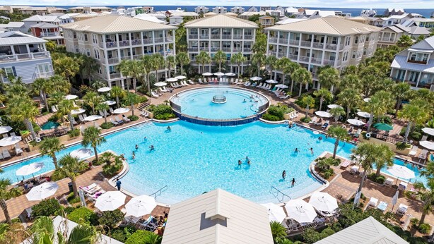 A resort with a central pool surrounded by palm trees and sun loungers, multiple buildings with balconies, and a view of the ocean.