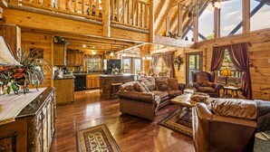 Een ruim houten blokhut interieur met een hoge nok, houten wanden, een gezellige zithoek met lederen meubels, een keuken en een groot raam dat natuurlijk licht binnenlaat.