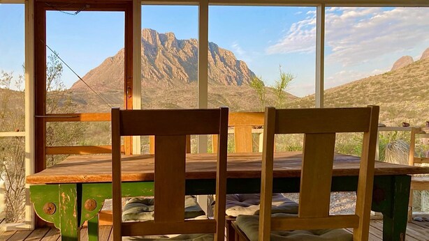 A wooden deck with two chairs and a table, overlooking a mountainous landscape through large windows. - Big Bend Getaway: Classic Adobe with Modern Flair (Terlingua)