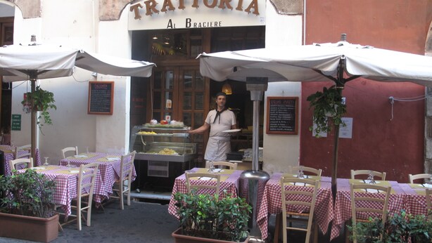 An outdoor dining area with tables covered in checkered tablecloths, a waiter serving food, and a sign reading "TRATTORIA Al Braciere". - SPECIAL OFFER - in the Heart of ancient Rome (Roma)