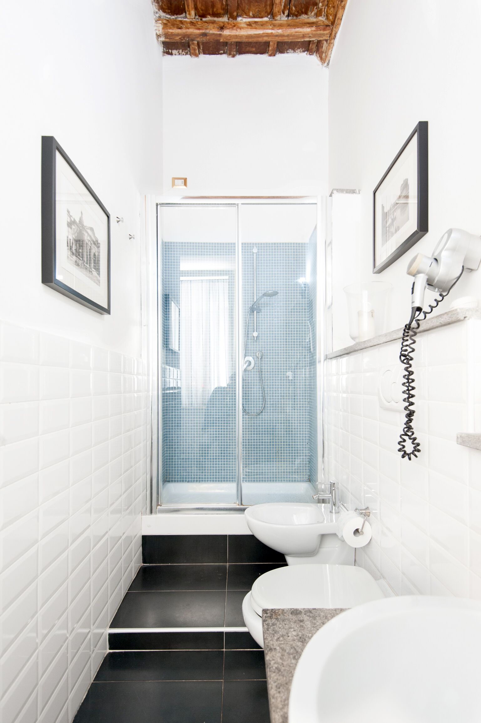 A bathroom with white tiled walls, a black tiled floor, a shower with a glass door, a toilet, and a sink with a hairdryer on the wall.