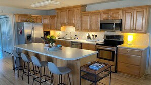 A modern kitchen with wooden cabinets, stainless steel appliances, a central island with bar stools, and a small side table with books.