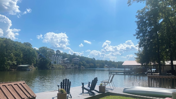 A lakeside deck with outdoor seating, a pool, and a view of a forested area with buildings in the background.