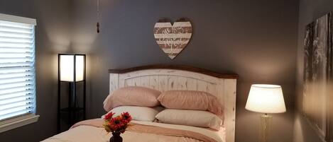 A bedroom with a bed, a ceiling fan, a heart-shaped wall decoration, a bedside table with a lamp, and a window with blinds.