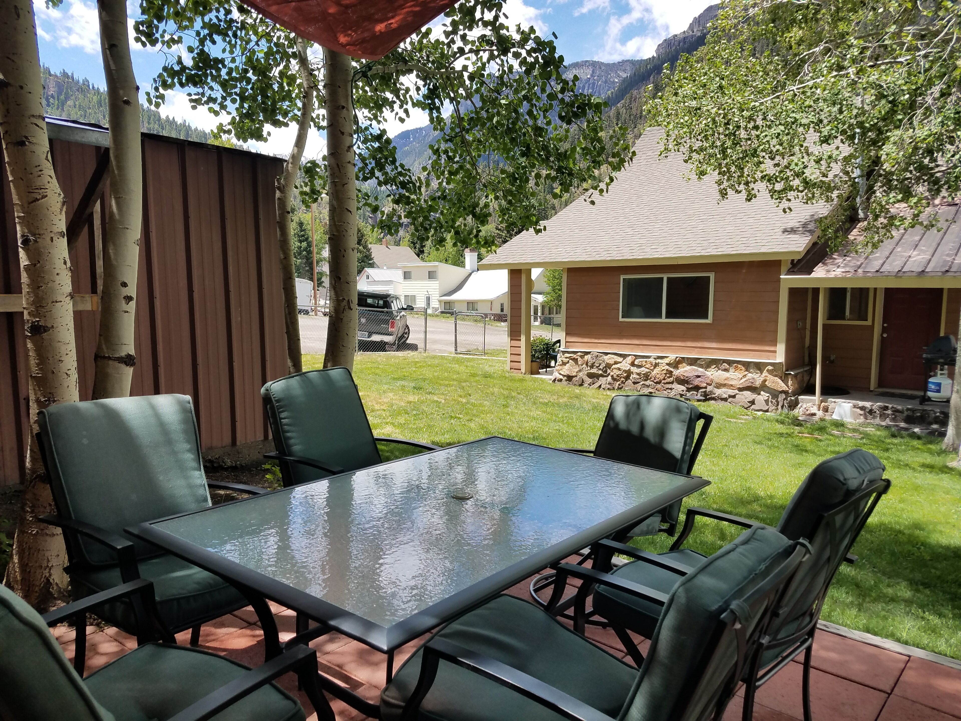 A patio with a glass table and chairs, surrounded by trees and a view of a mountain.
