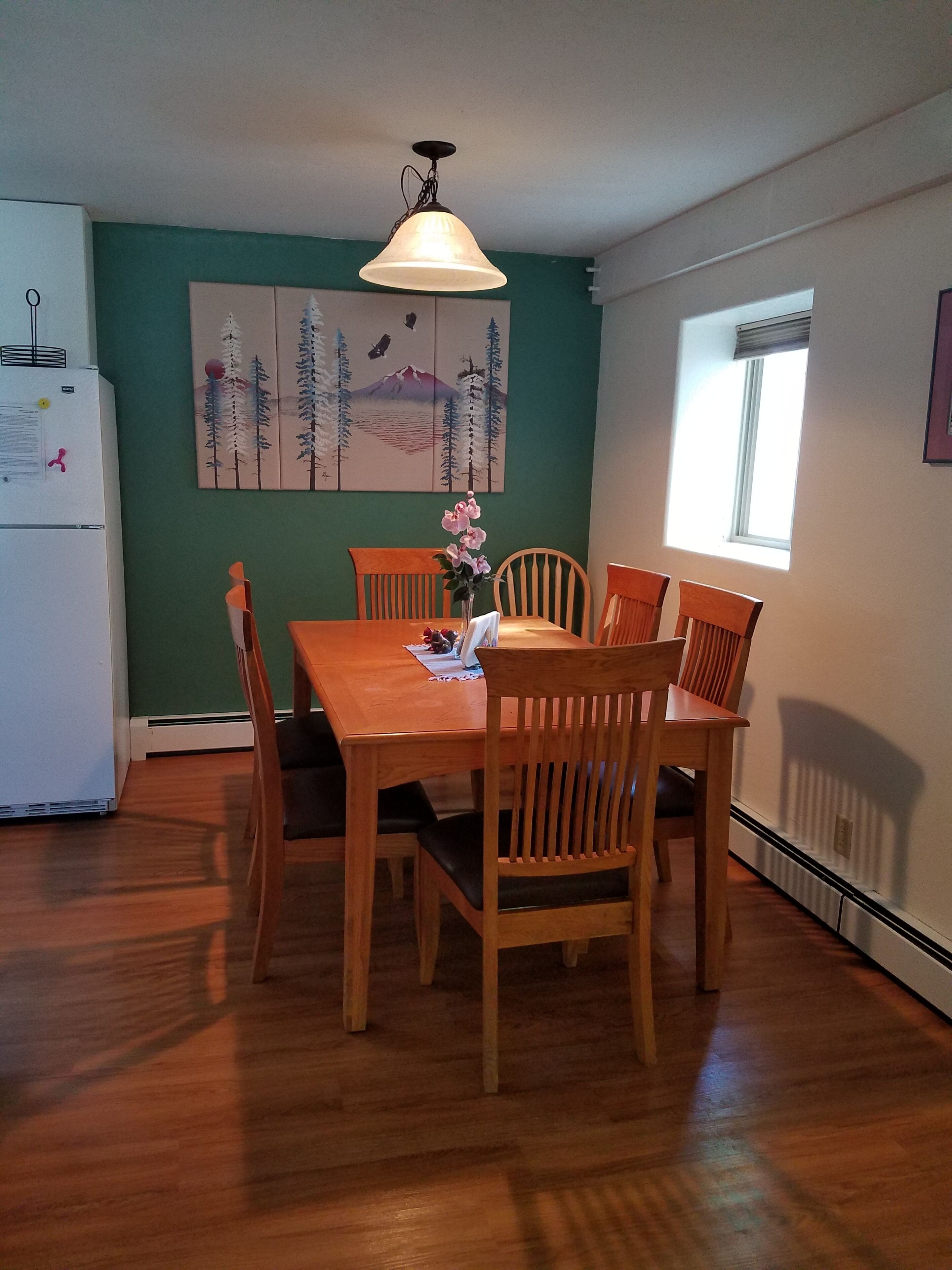 A dining area with a wooden table and chairs, a vase with flowers, a painting on the wall, and a window providing natural light.
