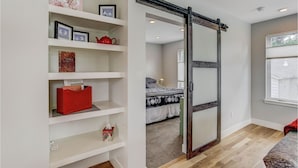 Un dormitorio con puerta corredera, una cama con colcha estampada, un baúl rojo y un estante con marcos de fotos y una tetera roja.