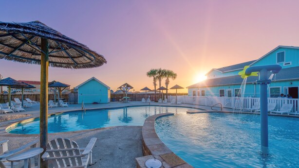 A pool area with lounge chairs, umbrellas, and a building with a blue roof. - Family-Friendly Townhome | Heated Pool, Patio & Beach Nearby (Corpus Christi)