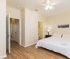 A clean, well-lit bedroom with a large bed, two bedside tables with lamps, a ceiling fan, and a door leading to another room.