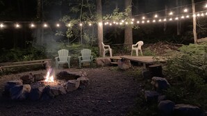 Un espace extérieur boisé, aménagé avec un foyer, deux fauteuils Adirondack et des guirlandes lumineuses.