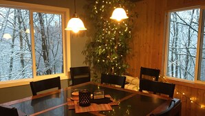 Un coin repas avec une table en verre entourée de chaises, une plante décorée de guirlandes lumineuses et des fenêtres donnant sur une forêt enneigée.