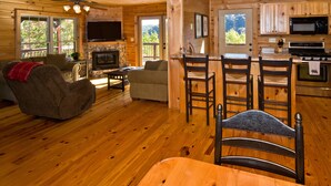 A cozy cabin interior with a stone fireplace, wooden furniture, and a kitchen area. - Beautiful Mountain Top Retreat Long Range Views, Hot Tub,Ping Pong,Horseshoe pit (Maggie Valley)
