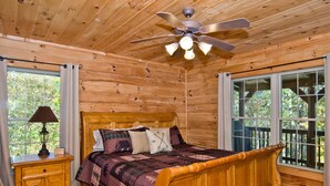 A wooden cabin bedroom with a bed, nightstand, lamp, ceiling fan, and large windows. - Beautiful Mountain Top Retreat Long Range Views, Hot Tub,Ping Pong,Horseshoe pit (Maggie Valley)