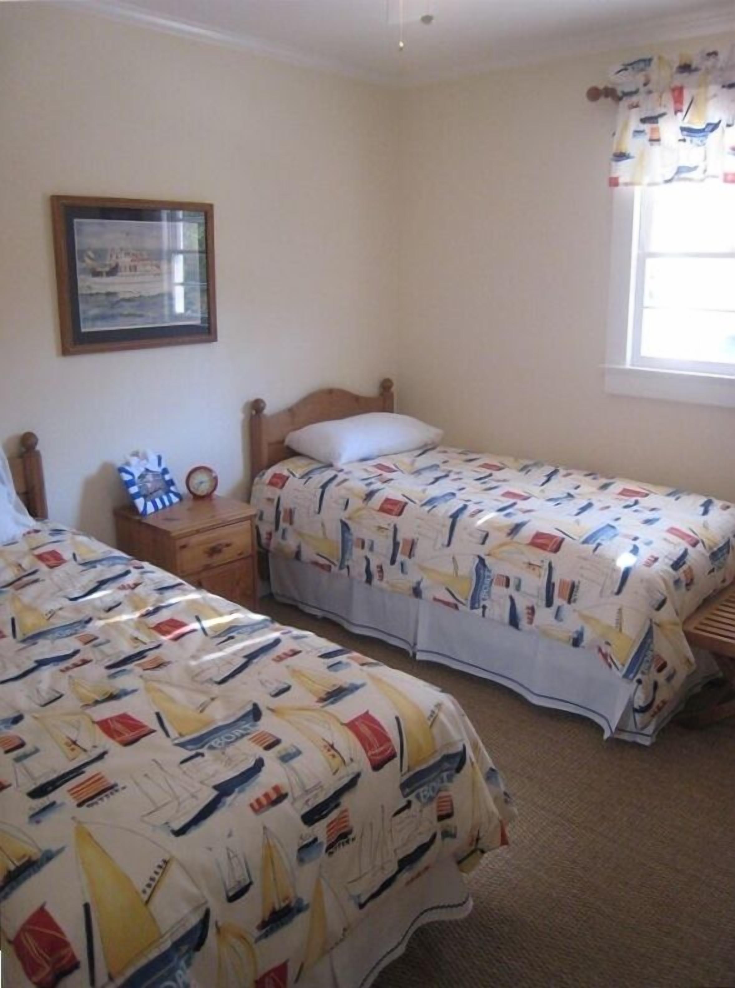 Un dormitorio con dos camas individuales, una mesita de noche, un cuadro enmarcado en la pared y una ventana con cortinas.
