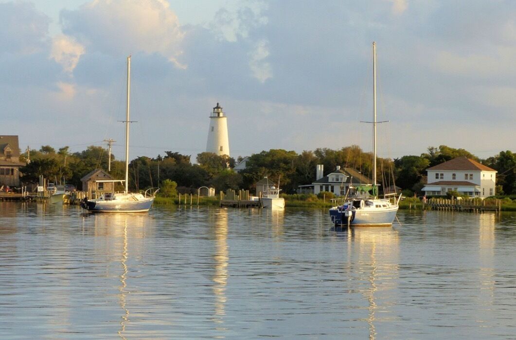 A calm waterfront with a lighthouse in the background, boats docked along the shore, and houses nearby.