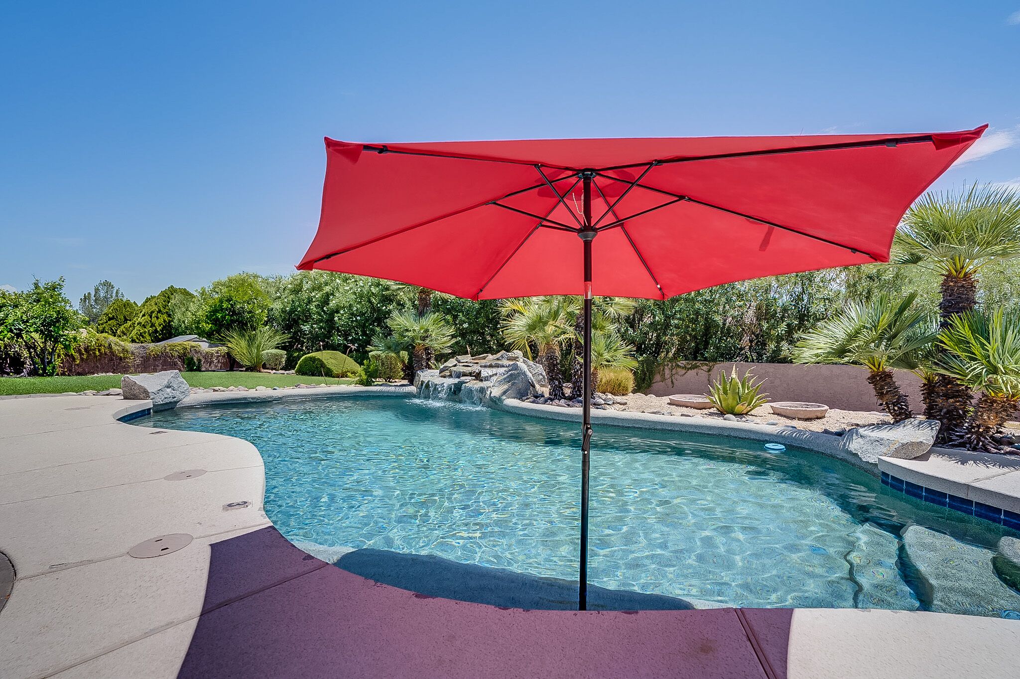 A swimming pool with a red umbrella, surrounded by landscaped greenery and a stone waterfall feature.