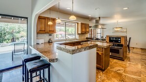 Una cocina moderna con encimeras de granito, armarios de madera y electrodomésticos de acero inoxidable. Hay taburetes de bar y una isla grande con un lavamanos.