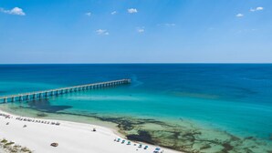 Une plage avec une jetée qui s’avance dans l’océan, une eau turquoise et limpide, et une rive sablonneuse parsemée de parasols.
