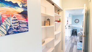 A hallway with white shelving units, a painting of a sunset with boats, and a kitchen area with stools and a clock.