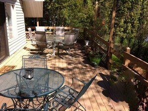 Une terrasse en bois avec une table et des chaises en verre, entourée d’une clôture en bois et de verdure.