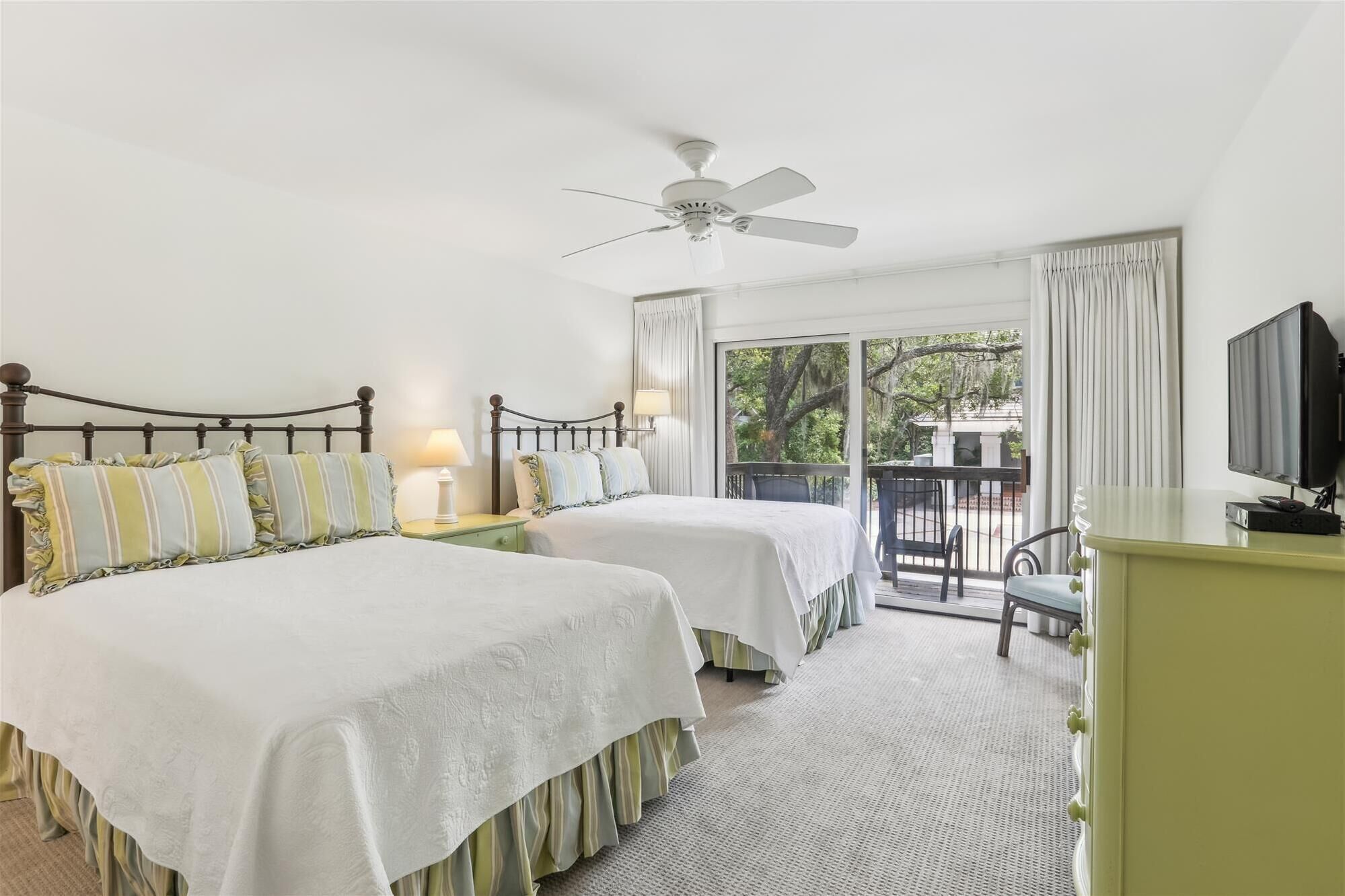 A hotel room with two beds, a ceiling fan, a television, and a balcony with a view of trees.