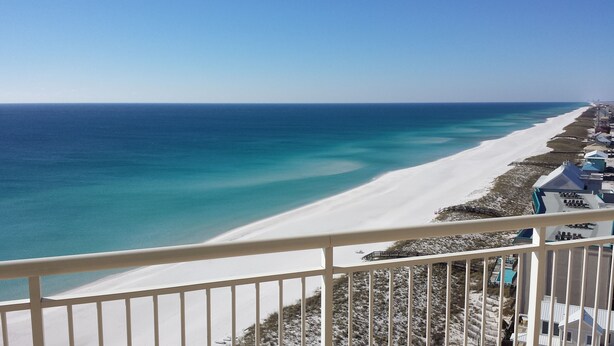 A beach view with a white sandy shore, clear blue water, and a row of buildings with a blue roof along the coastline. - Spacious Belle Mer 3BR -- 2 Gulf Front Masters -- All with Private Baths (Navarre Beach)