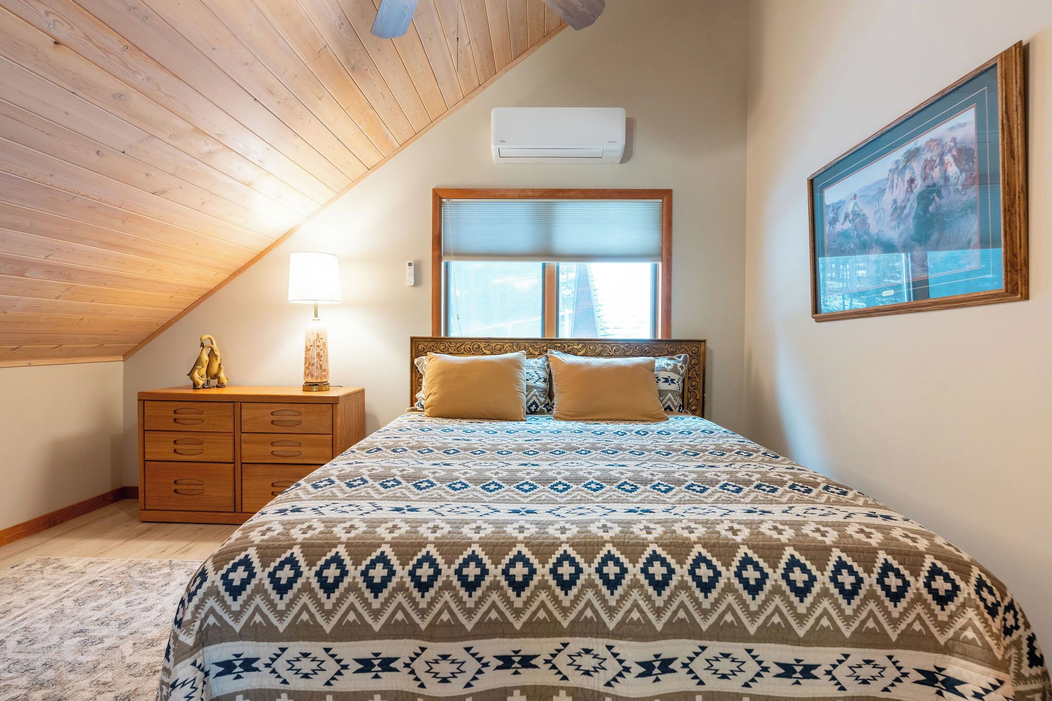 Een slaapkamer met schuine wand, een bed met beddengoed met patroon, een houten nachtkastje, een lamp, een ingelijst schilderij aan de muur en een airconditioner.