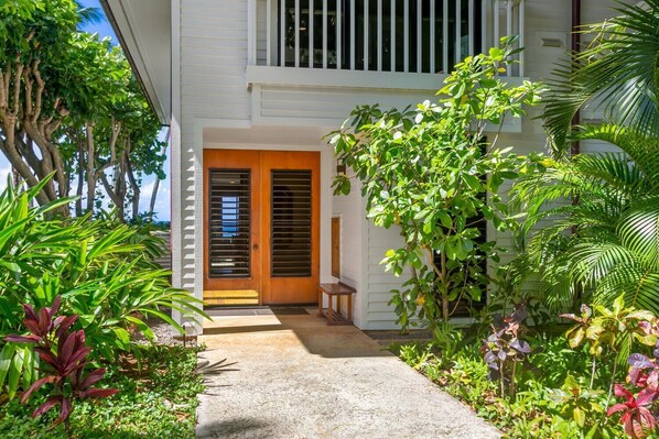 A house with a wooden door and a small porch. There are various plants and trees around the house. - Poipu Kapili 6 by Alekona Kauai: Oceanfront Corner Condo w AC (Koloa)