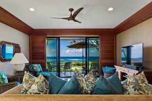 A room with a large TV, a ceiling fan, and a view of the ocean through a sliding glass door. - Poipu Kapili 6 by Alekona Kauai: Oceanfront Corner Condo w AC (Koloa)