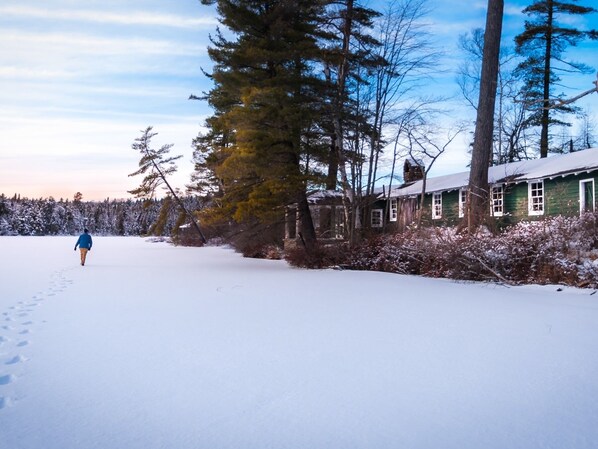 A snow-covered landscape with a person walking away from the camera, a green building with white trim, and a forest in the background.