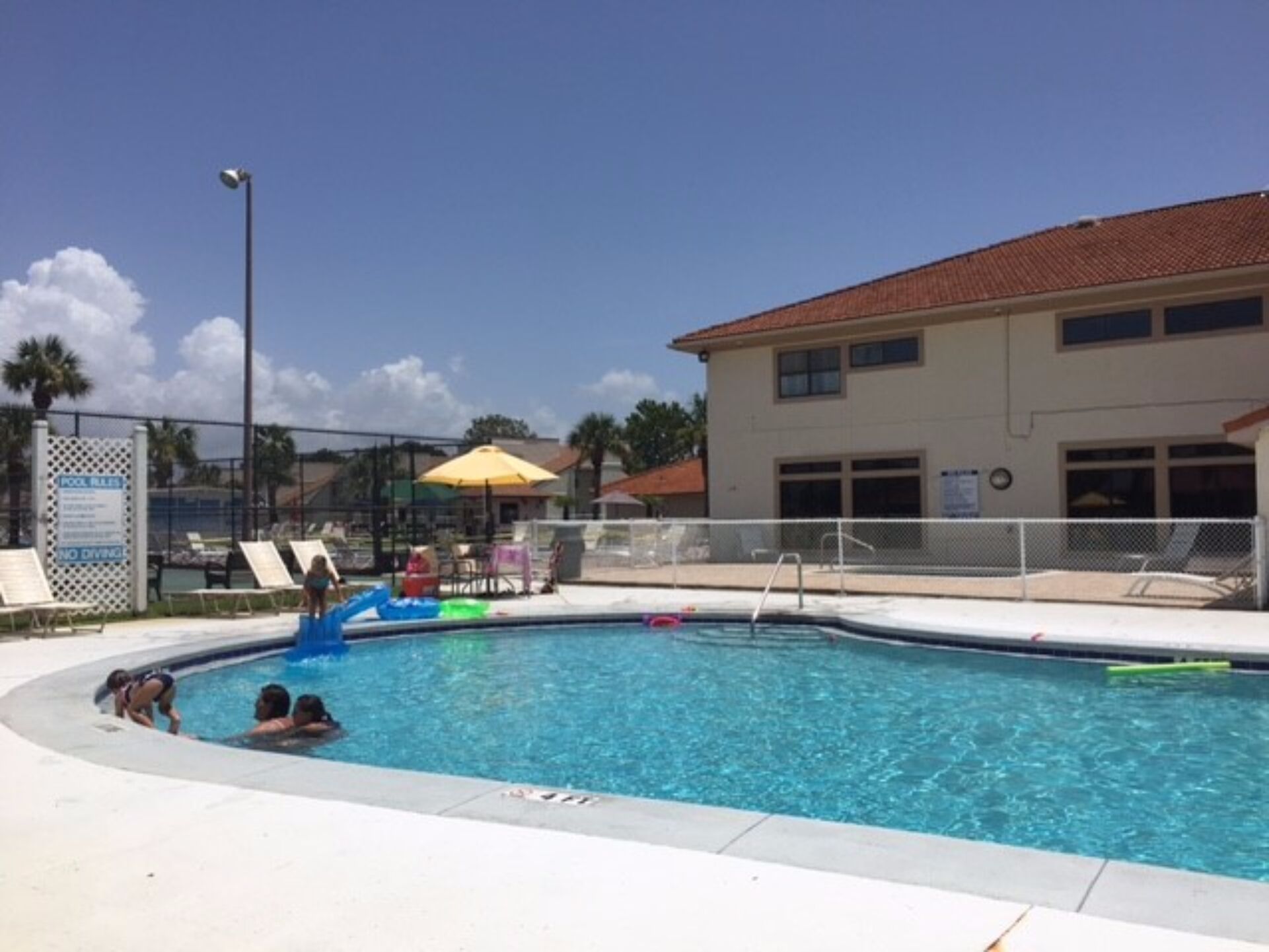 A swimming pool with a fenced area, lounge chairs, and a building with a red roof in the background.