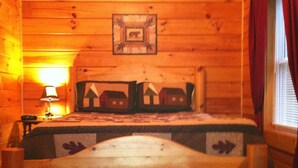 A wooden cabin bedroom with a bed, a nightstand, a lamp, and a window with curtains.