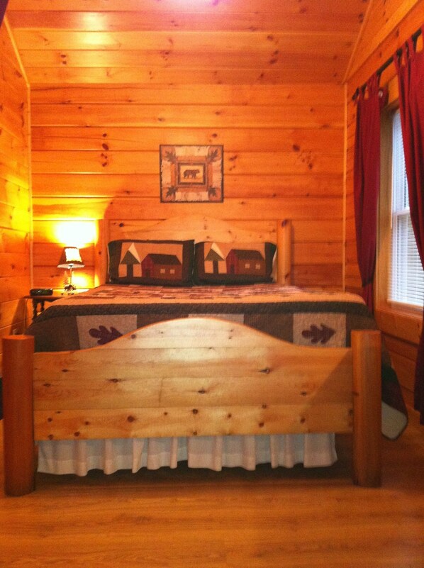 Une chambre à coucher dans une cabane en bois, avec un lit, une table de chevet, une lampe et une fenêtre avec des rideaux.