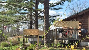 A cabin with a deck, surrounded by trees and a grassy area with yellow flowers.