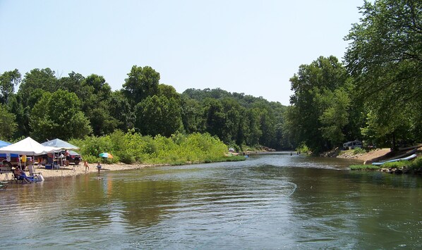 Un río con zona de camping en sus orillas, incluyendo tiendas de campaña y una canoa.