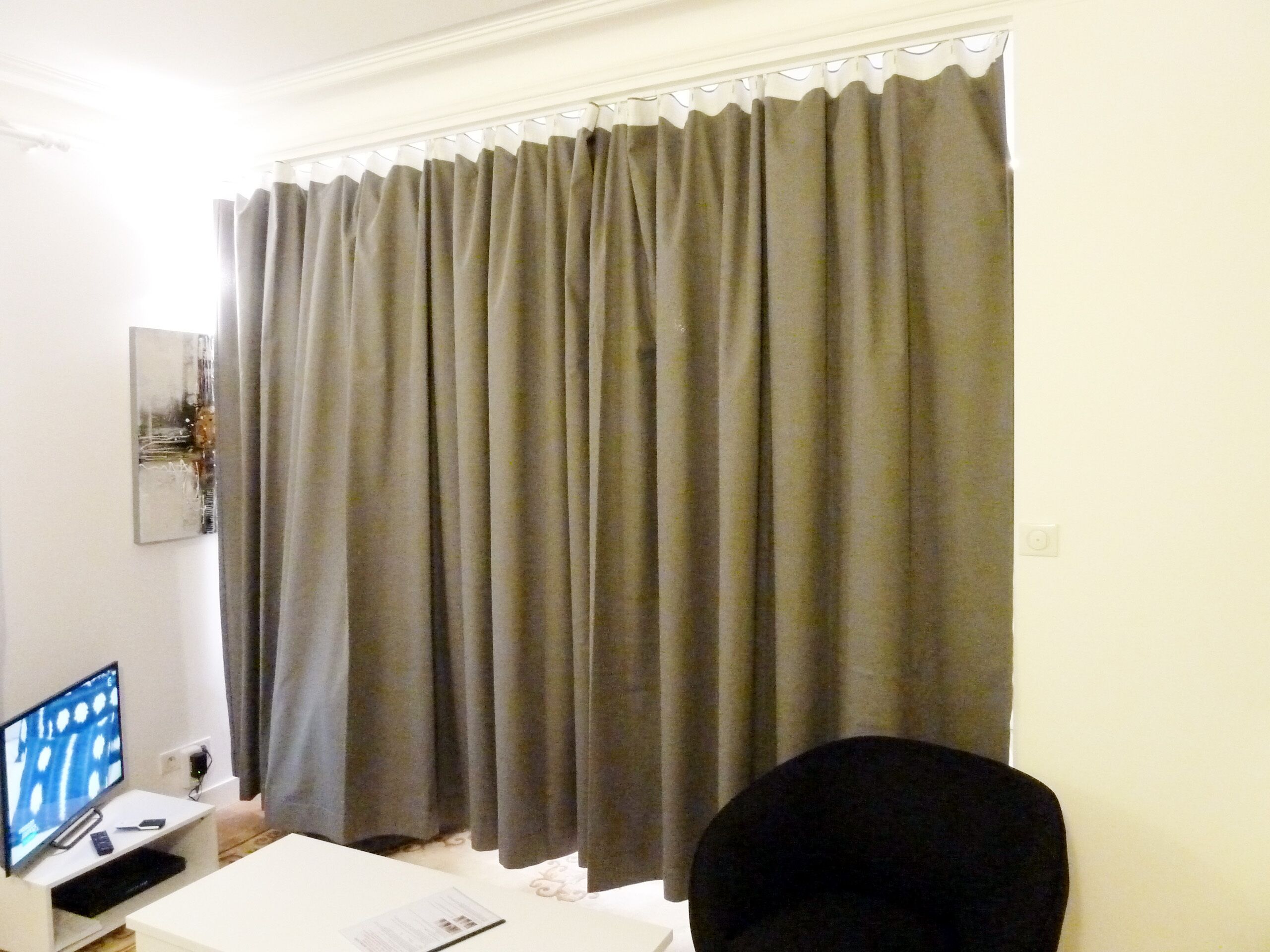 Een kamer met een groot grijswas gordijn, een wit bureau met een computer, een zwarte stoel en een klein kunstwerk aan de muur.