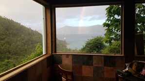 A room with a large window offering a view of a lush landscape, a rainbow, and a distant island. - Gorgeous View, Beach Access, Swimming Pool, Kayak, Quiet Privacy - the East End (St. John)