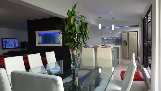 A modern living room with a glass dining table, white sofas, a TV, and a kitchen in the background.