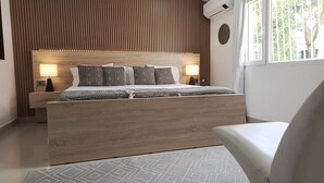 A modern bedroom with a wooden headboard, a bed with patterned bedding, bedside lamps, and a view of greenery outside.