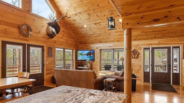 Interior de una cabaña de troncos con comedor, sala de estar con chimenea y televisor.