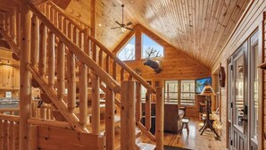 Interior de una cabaña de madera con escalera, comedor y un ventanal grande.