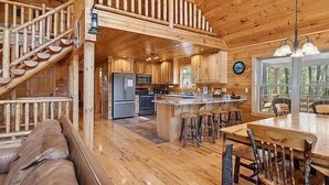 Interior de una cabaña con escalera de madera, cocina, comedor con mesa y sillas de madera, y sala de estar con sofá.