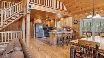L’intérieur de la cabane comprend un escalier en bois, un coin cuisine, un espace repas avec une table et des chaises en bois, ainsi qu’un coin salon avec un canapé.