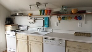 A kitchen with a stove, sink, and dishwasher, wooden cabinets, and a shelf with colorful mugs and dishes. - Adorable Loft Apartment (Wildwood Crest)