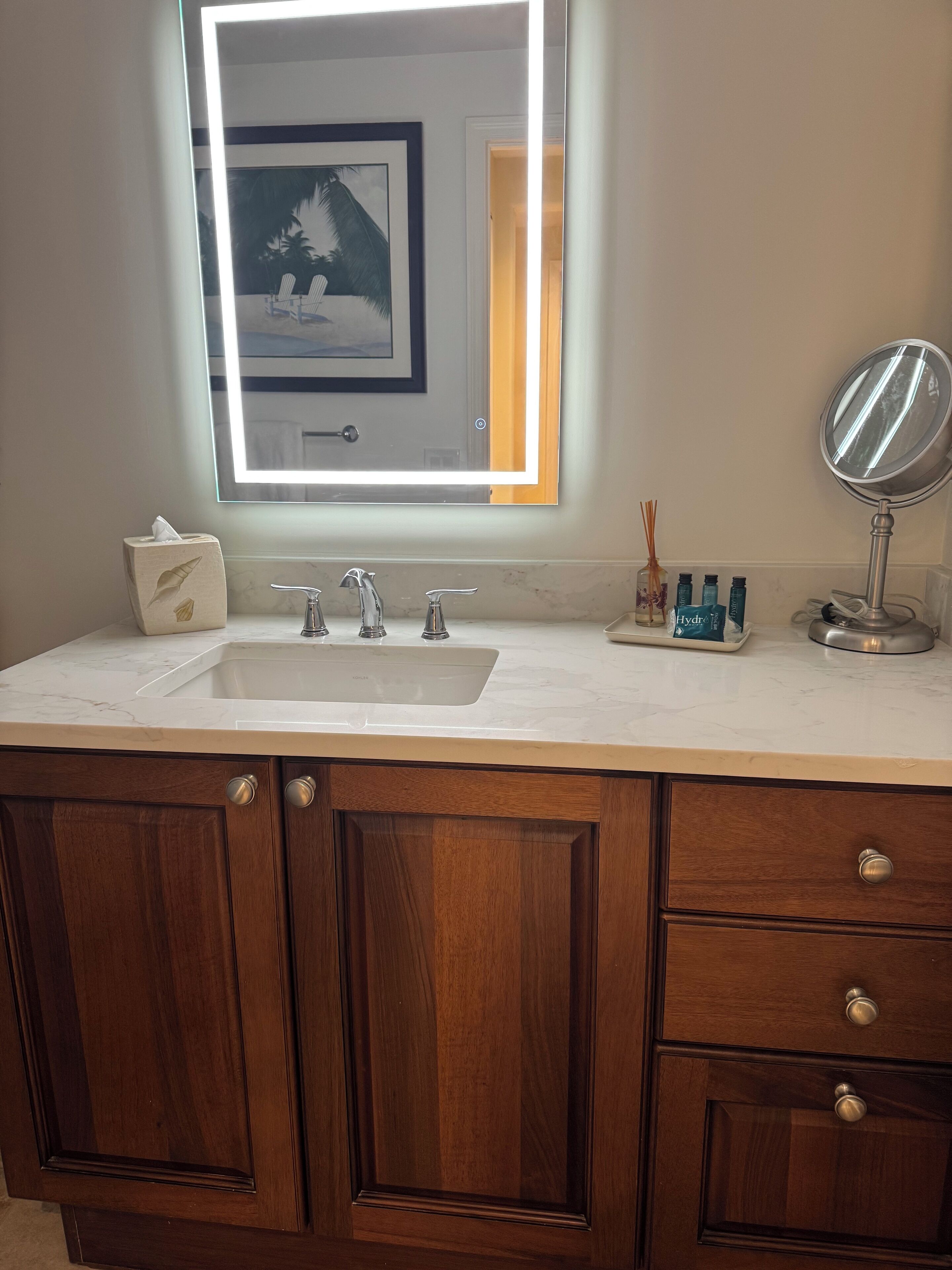 A bathroom with a marble countertop, a sink with a modern faucet, a wooden vanity with drawers, a framed picture above the mirror, and a small bottle of hand soap on the counter.