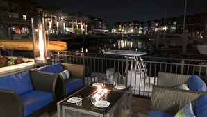 Un área de estar al aire libre con una mesa y sillas, una lámpara de mesa iluminada y vista hacia un puerto deportivo con barcos y edificios al fondo.