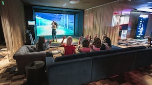 Un grupo de personas observando una presentación en una sala con una pantalla grande, asientos cómodos y suelo a cuadros.