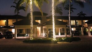 Un hotel con dos palmeras iluminadas, una zona de estacionamiento techada y un edificio con varios balcones.