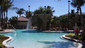 Piscina con caída de agua, rodeada de palmeras y un paseo enlosetado.