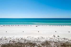 Una playa extensa con hileras de sombrillas azules y sillas de playa blancas, que se extiende hasta un mar tranquilo bajo un cielo despejado.
